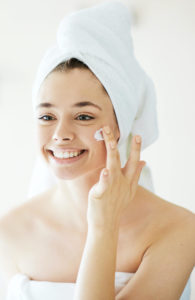 a smiling young woman in a towel applying skin moisturizer on her face